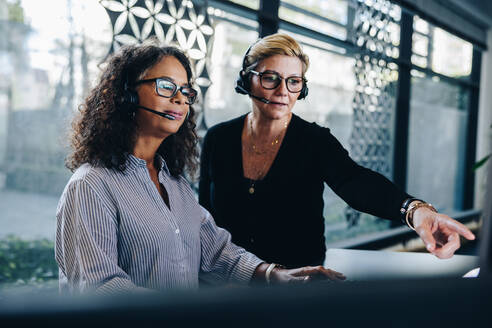 Zwei Geschäftsfrauen mit Headset, die gemeinsam am Computer arbeiten. Ein Manager zeigt auf den Computerbildschirm und diskutiert etwas mit einem Kollegen, der am Schreibtisch arbeitet. - JLPSF05286