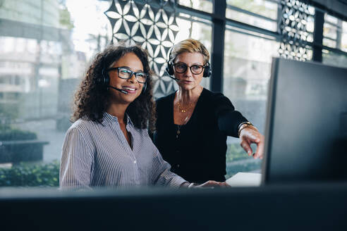 Zwei Geschäftsfrauen, die zusammenarbeiten, während sie einen Computer benutzen und im Büro arbeiten. Ein Manager hilft einer Kollegin bei der Arbeit am Computer. Beide tragen ein Headset und arbeiten in einem Call Center. - JLPSF05290