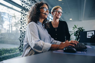 Lächelnde Frau mit Headset, die an einem Computer arbeitet, mit einem Manager, der daneben steht. Zwei weibliche Geschäftsleute in einem Callcenter. - JLPSF05291