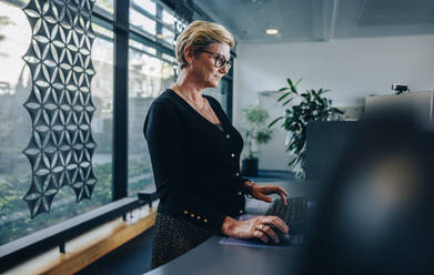 Senior businesswoman working at standing desk. Woman employee working on desktop computer at ergonomic standing desk. - JLPSF05292