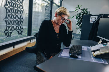 Senior businesswoman looking tired sitting at her desk. Female professional feeling stressed while working in office. - JLPSF05296