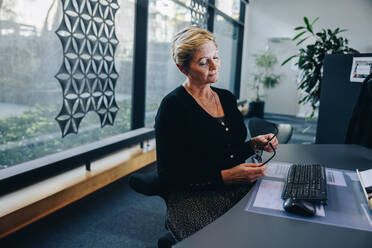 Mature businesswoman taking a break from work. Female professional holding her eyeglasses and sitting idle at her desk. - JLPSF05297