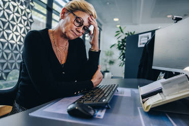 Weibliche Fachkraft hat Stress bei der Arbeit im Büro. Ältere Geschäftsfrau sitzt an ihrem Schreibtisch und sieht müde aus. - JLPSF05302