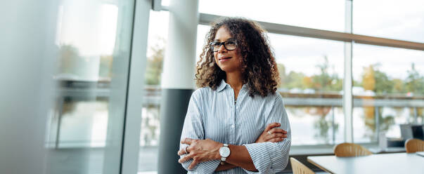 Woman entrepreneur standing beside a window in office. Mature woman standing in office with arms crossed looking outside window. - JLPSF05350