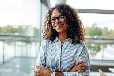 Portrait of female professional with curly hair wearing eyeglasses. . Smiling businesswoman with her arms crossed - JLPSF05352