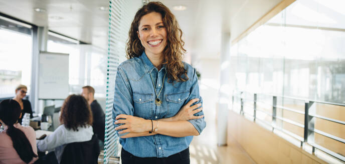 Portrait of a successful businesswoman standing outside office boardroom with colleagues discussing in background. Happy looking female professional with her arms crossed. - JLPSF05387