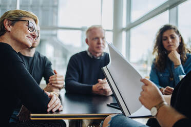 Ältere Geschäftsfrau, die auf die Vertragsdiskussion im Sitzungssaal achtet, Geschäftsleute, die an einem Konferenztisch sitzen und einen Kollegen, der ein Papier liest. - JLPSF05392