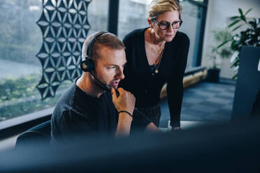 Businessman wearing headset looking at computer and thinking with female manager besides. Customer service representative at work with manager. - JLPSF05410