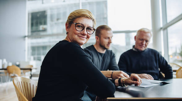 Happy senior businesswoman sitting at a meeting in boardroom. Female professional with coworkers in conference meeting. - JLPSF05412