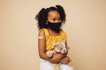 Small girl wearing protective face mask sitting with her teddy bear. Girl child with face mask on brown background after getting vaccination. - JLPSF05451