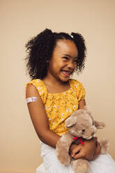 Cute girl with bandage on arm after getting a vaccine smiling. Girl holding her teddy bear looking away and smiling after vaccination. - JLPSF05452