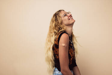 Junge Frau lachend auf braunem Hintergrund mit einem Verband am Arm nach der Impfung. Frau fühlt sich nach der Impfung. - JLPSF05461