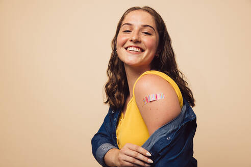 Portrait of a female smiling after getting a vaccine. Woman holding down her shirt sleeve and showing her arm with bandage after receiving vaccination. - JLPSF05470