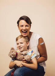 Cheerful mother and son after vaccination on brown background. Woman holding her son with a teddybear. - JLPSF05477