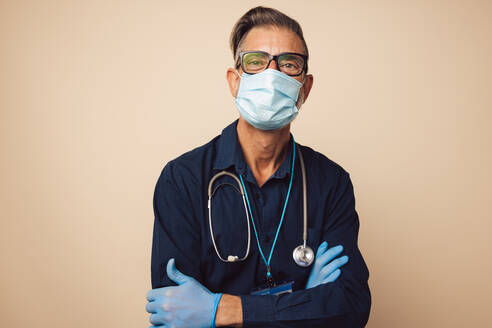 Porträt eines männlichen Arztes mit Schutzmaske und Handschuhen, der mit verschränkten Armen im Gesundheitswesen arbeitet. - JLPSF05484