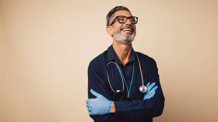 Porträt eines fröhlichen Allgemeinmediziners mit Stethoskop und Schutzhandschuhen, der mit verschränkten Armen dasteht und lächelt. - JLPSF05485