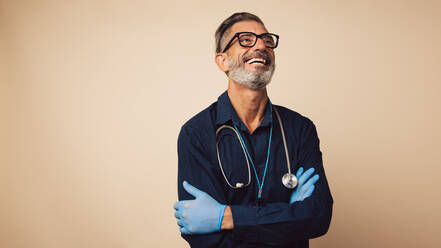 Portrait of a cheerful general practitioner with stethoscope wearing protective hand gloves. Doctor standing with his arms crossed looking away and smiling. - JLPSF05485