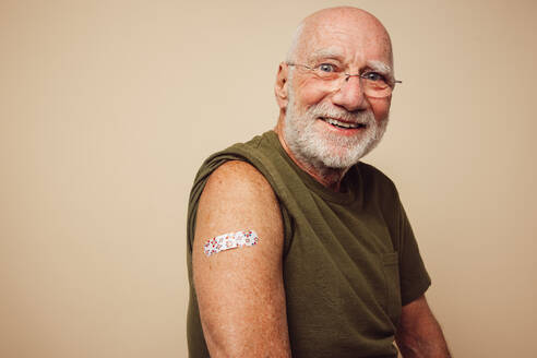 Portrait of senior man smiling after getting vaccine. Mature man showing his arm with bandage after receiving vaccination. - JLPSF05494