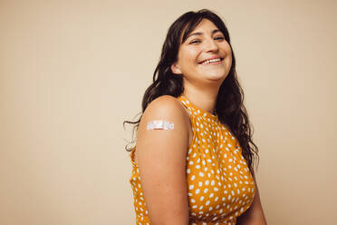 Beautiful mature with bandage on her arm after she received vaccination. Woman smiling on brown background feeling happy to get inoculated. - JLPSF05497