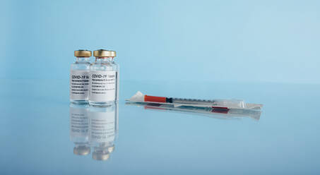 Covid-19-Impfstofffläschchen und Spritze auf reflektierender Oberfläche. Corona-Virus-Impfflaschen mit Injektion auf blauem Hintergrund. - JLPSF05512