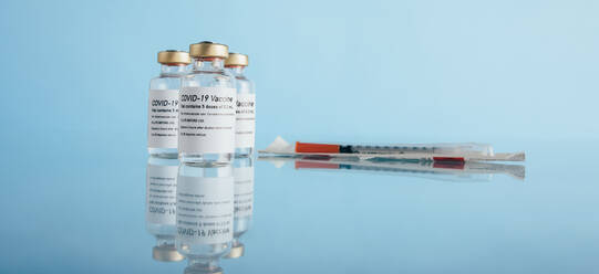 Covid-19 vaccine vials and syringe on reflective surface. Corona virus vaccination bottles with injection on blue background. - JLPSF05514