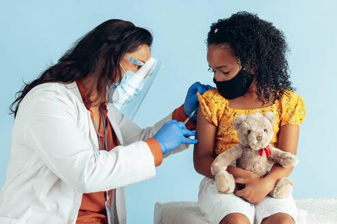 Female doctor wearing protective face mask, gloves and shield injecting vaccine into arm of girl in mask during pandemic. Small girl holding her teddybear getting covid-19 vaccine from a physician. - JLPSF05528