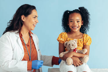 Happy female doctor with a small girl holding teddy bear smiling. Pediatric with girl patient on blue background. - JLPSF05529