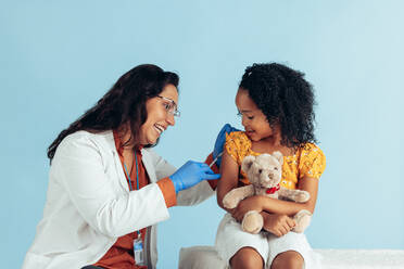 Lächelnder Arzt, der einem kleinen Mädchen mit Teddybär einen Impfstoff verabreicht. Mädchen, das in einer Klinik eine Grippeimpfung am Arm erhält. - JLPSF05531