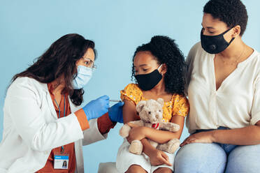 Ein Arzt mit Gesichtsmaske, Handschuhen und Schutzschild verabreicht einem Mädchen mit Maske einen Impfstoff. Ein kleines Mädchen, das seinen Teddybären hält, wird von einem Kinderarzt gegen Covid-19 geimpft. - JLPSF05536