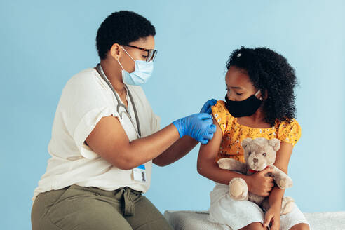 Female doctor putting bandage on arm of a girl after giving vaccine. Doctor and girl wearing face masks during vaccination. - JLPSF05541