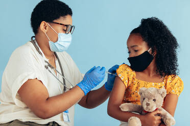 Kinderarzt, der einem Mädchen eine Spritze in den Arm gibt. Arzt, der einem Mädchen mit Teddybär in der Klinik eine Impfung verabreicht. Beide tragen Gesichtsschutzmasken. - JLPSF05542