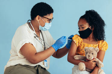 Ärztin, die einem Mädchen eine Impfung verabreicht. Medizinische Fachkraft und kleines Mädchen tragen bei der Impfung gegen das Coronavirus Gesichtsmasken. - JLPSF05543