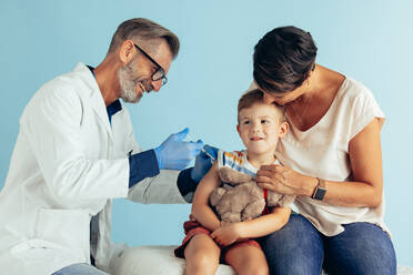 Lächelnder reifer Arzt, der einem Jungen eine Impfung verabreicht. Ein Junge wird geimpft, während er mit seiner Mutter sitzt. - JLPSF05568