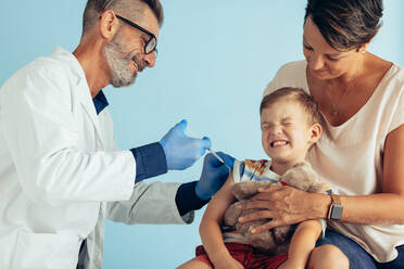 Ein Arzt gibt einem Jungen, der sein Auge vor Schmerzen schließt, eine Injektion in den Arm. Eine männliche medizinische Fachkraft gibt einem Kind, das mit seiner Mutter zusammensitzt, einen Impfstoff. - JLPSF05571