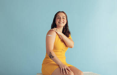Pretty woman showing her arm after getting vaccine. Female with band-aid on her arm after receiving flu shot on blue background. - JLPSF05579