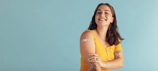 Schöne Frau sieht glücklich aus, nachdem sie geimpft wurde. Frau mit Pflaster auf dem Arm, nachdem sie geimpft wurde, sitzt auf blauem Hintergrund. - JLPSF05582