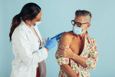 Female doctor giving coronavirus vaccine to mature man. Medical professional and mid adult man wearing protective face masks during covid-19 vaccination. - JLPSF05593