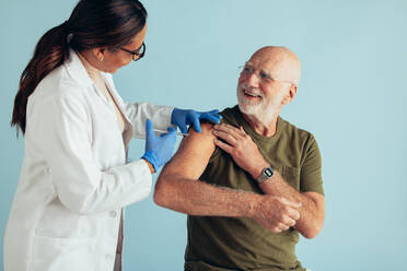 Senior man getting flu vaccine injection by a female medical professional. Doctor giving vaccination to elderly man on blue background. - JLPSF05600