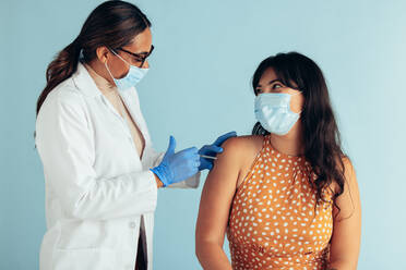 Female doctor and woman patient wearing face masks during vaccination. Medical professional giving vaccine to female against blue background. - JLPSF05605