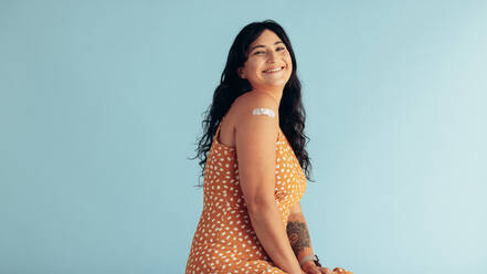 Smiling woman after getting vaccine on blue background. Happy woman showing her arm with bandage after receiving vaccination. - JLPSF05608