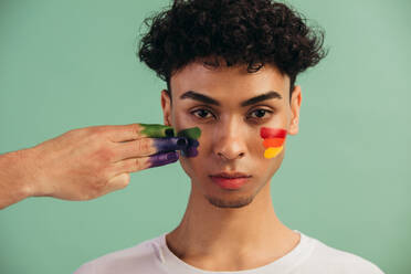 Closeup of a hand painting LGTB flag colors on man's face. Transgender male with gay pride flag painted on his cheeks. - JLPSF05692