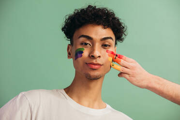 Portrait of young man with face painted with rainbow flag of gay pride. Hand painting lgbt rainbow color on a man's face. - JLPSF05695