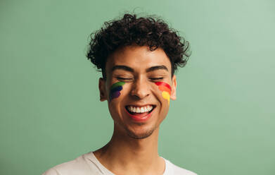 Close-up of a cheerful young man with pride flag painted on face. Gay man with rainbow face paint smiling against pastel green background. - JLPSF05698