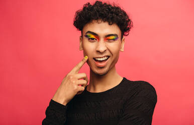 Gay man wearing multicolored shadows on the eyelids winking at camera. Happy transgender male winking an eye against red background. - JLPSF05706