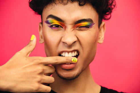 Close-up of a transgender male biting his finger and winking an eye. Gender fluid man wearing rainbow colored eye shadow and smiley face fingernail. - JLPSF05708