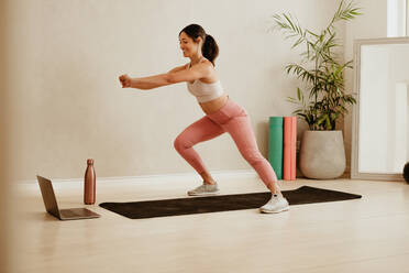 Woman doing lunges workout watching online class. Female in fitness wear teaching exercise to students online with her laptop. - JLPSF05727