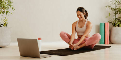 Frau beim Sport auf Yogamatte sitzend mit Laptop vor sich. Lächelnde Frau in Sportkleidung beim Trainieren und Beobachten eines Online-Fitnesskurses. - JLPSF05733