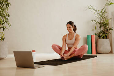 Woman sitting on yoga man and doing stretching workout while walking online video on laptop. Sporty woman doing exercises at home while watching online training from her laptop. - JLPSF05734