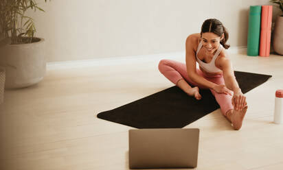 Female fitness trainer teaching leg stretching exercises online on laptop. Woman working out in front of her laptop in fitness studio. - JLPSF05739