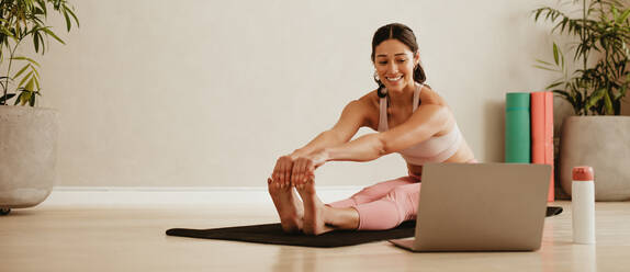Fit woman doing yoga exercise while watching tutorial on laptop at home. Attractive woman exercising on a mat and watching instructional videos on laptop. - JLPSF05740